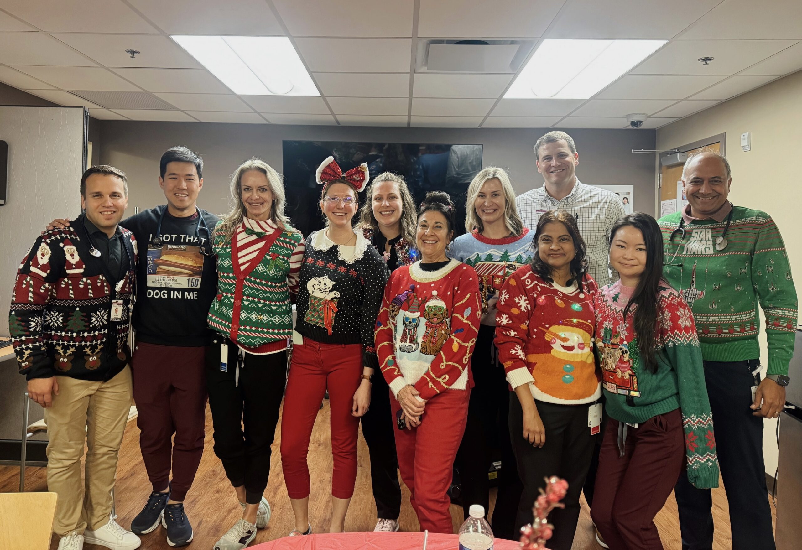 A group of eleven healthcare professionals standing together in a room, smiling and wearing festive "ugly" Christmas sweaters. Several people are wearing stethoscopes around their necks. The sweaters feature various designs, including Santa Claus, reindeer, snowmen, Star Wars themes, and one humorous shirt referencing a Costco hot dog. The group is posed in two rows against a neutral-colored wall under bright office lighting.