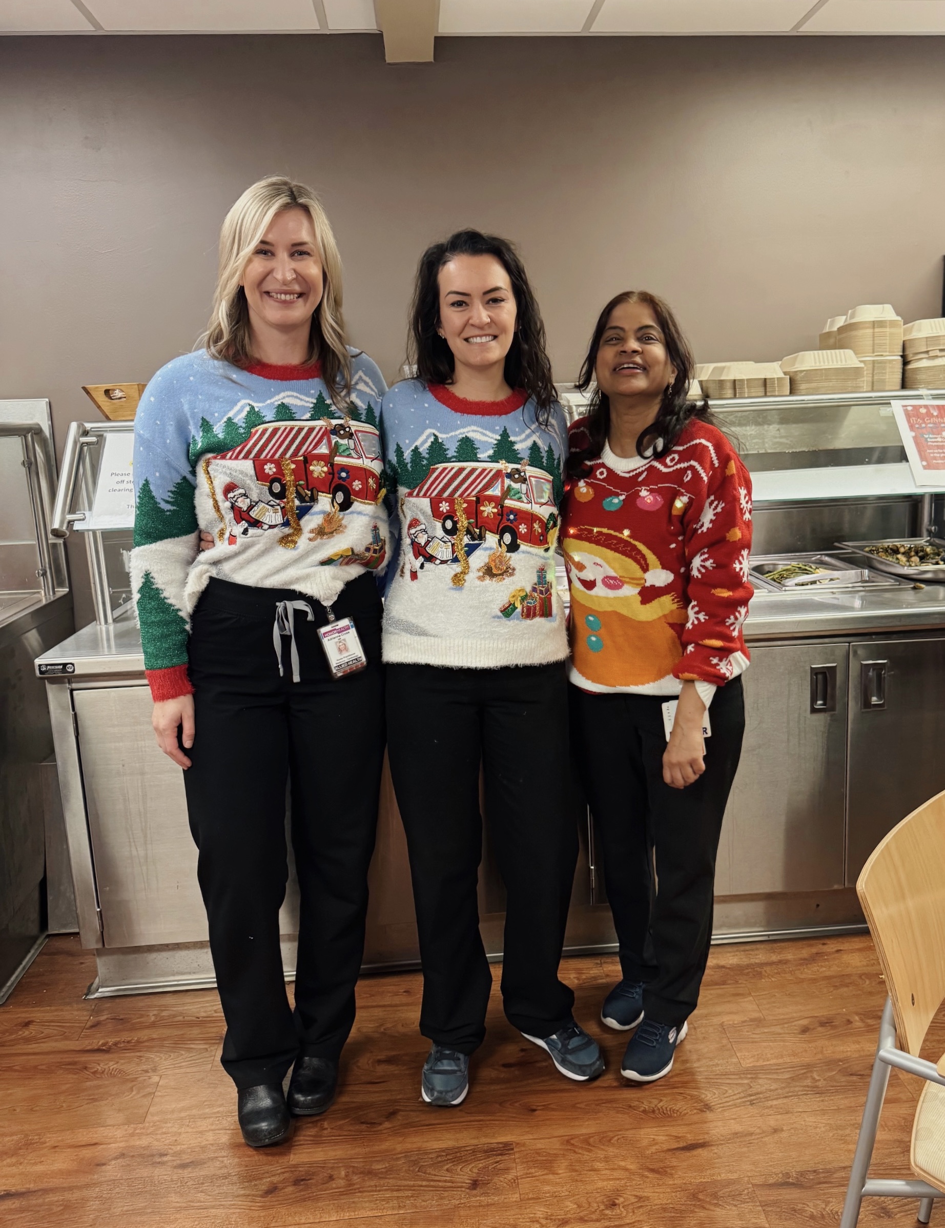 Three women standing together in a physicians lounge setting, smiling and wearing holiday sweaters. Two of the women wear matching light blue sweaters featuring a red camping van in a snowy forest scene. The third woman wears a red sweater with a large, smiling snowman and colorful lightbulb patterns. They are posed in front of a stainless steel food service counter.