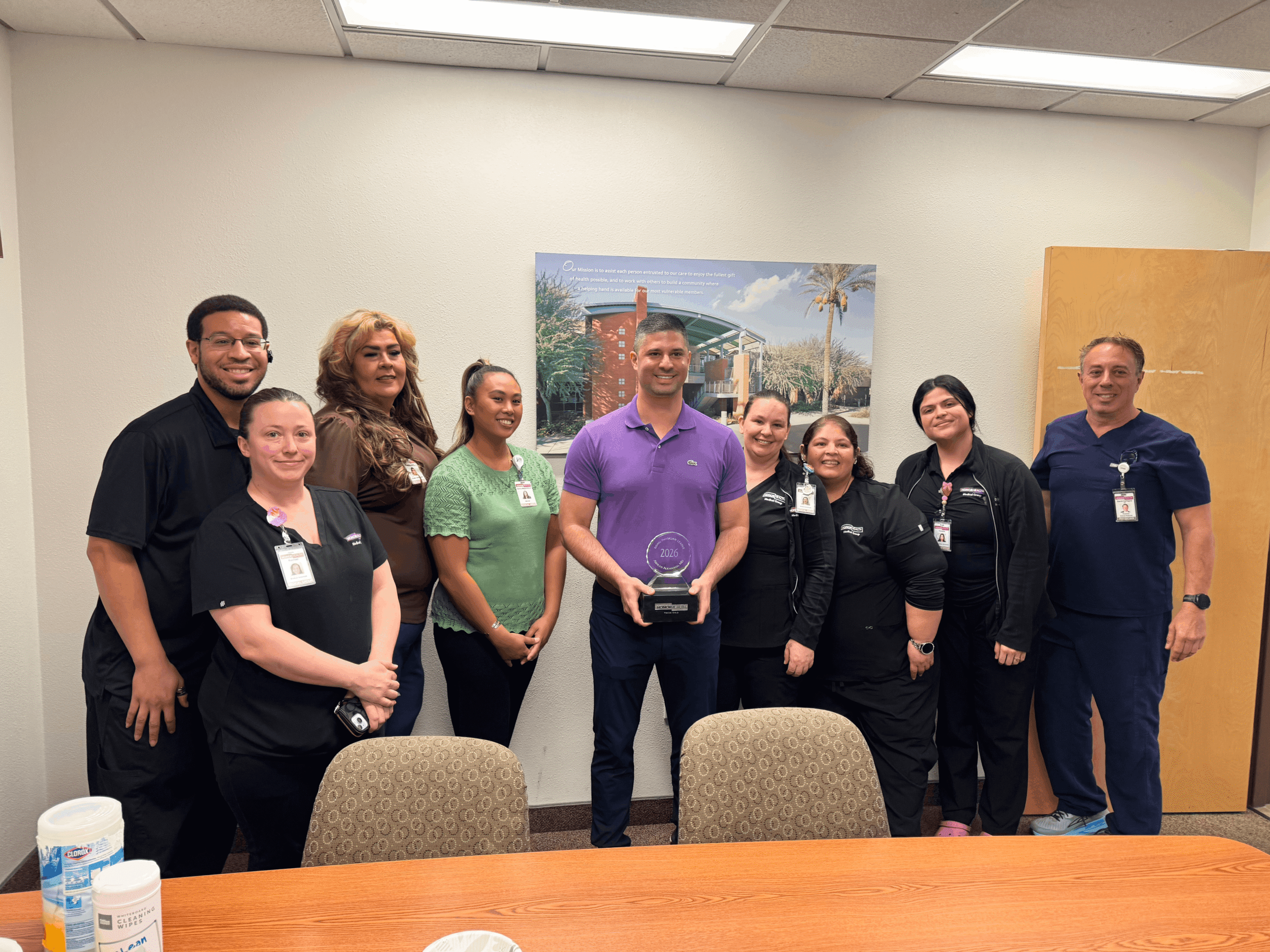 A group of nine HonorHealth team members posed in an office setting. Dr. Alkhamisi in a purple polo shirt stands in the center holding a circular glass award. Several colleagues in scrubs and professional attire stand beside him, all smiling.