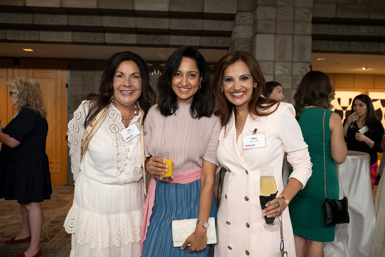Three women smiling and holding beverages at an indoor networking event. They are wearing name tags with the WPLC logo.