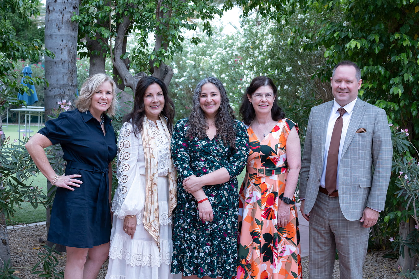 Four women and one man standing together and smiling outside in front of green trees and bushes at the WPLC Happiness Brunch.