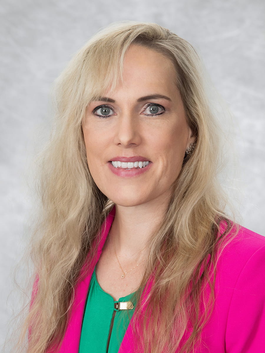 Professional headshot of Dr. Tabitha Moe wearing a bright pink blazer and a green top against a light gray background.